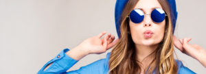 Chic young redheaded woman pursing her lips, wearing blue sunglasses, hat and blouse