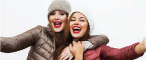 Two best girlfriends in winter coats and knit hats laughing, taking a selfie.