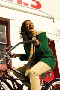 Laughing woman wearing a green sweater and gold/gray scarf, riding a bicycle.