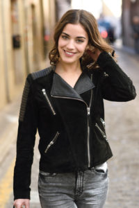 Young smiling brunette woman wearing a black suede jacket, walking down the street.